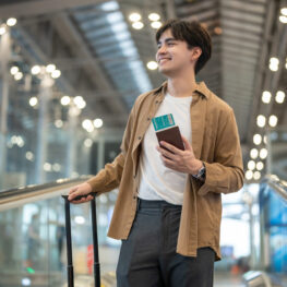 Asian young man passenger walking in airport terminal to boarding gate. Attractive handsome male tourist feeling happy and excited for going travel abroad by airplane alone for holiday vacation trip.