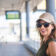 Confident woman in sunglasses and cap smiles at camera in a bright modern transit station, ready for urban commute or travel with relaxed, stylish, optimistic energy